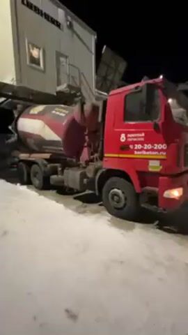 A red concrete mixer truck is parked in a snow-covered area, next to a large building bearing the name "Liebherr." The truck is facing right, with its headlights on. The concrete mixer drum is painted with stripes and lettering in Russian. The side of the truck displays a phone number and the website "beribeton.ru." The scene is set at night, with artificial lighting illuminating the truck and surrounding area. The camera is static, offering a close view of the truck and the building beside it.