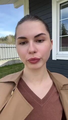 A young woman stands outside near a dark gray house on a sunny day. She is facing the camera, wearing a brown V-neck sweater, a light brown trench coat slung over her shoulders, and white pants. She has her hair pulled back, and after an initial neutral expression, she smiles at the camera. The background includes a green lawn, a metal fence, and trees in the distance, visible under a clear blue sky.