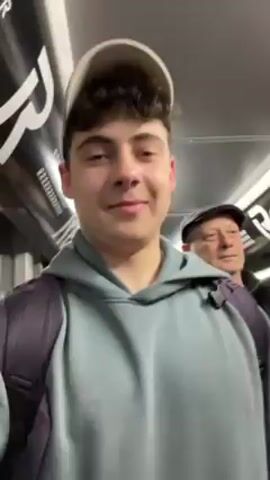 A young man with curly brown hair and a white baseball cap is recording a selfie video on a crowded train. He wears a light blue hoodie and a backpack. Behind him, an older man in a black jacket and a black flat cap looks forward. Many other passengers are visible, including a woman in a green jacket. The train car has black and white advertisements on the upper walls. People are standing and moving. The lighting is bright.