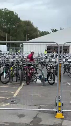 The video shows a cyclist in a red shirt, black shorts, and a white helmet, pushing their bike through a crowded bicycle parking area. The parking area is filled with numerous bicycles secured in rows of racks. In the background, a white tent-like structure is visible, suggesting a possibly organized event or competition. The day appears overcast. A second person in a blue shirt and black shorts with sunglasses walks in front of the camera. The cyclist appears to be heading away from the tent and towards the street or another section of the event.