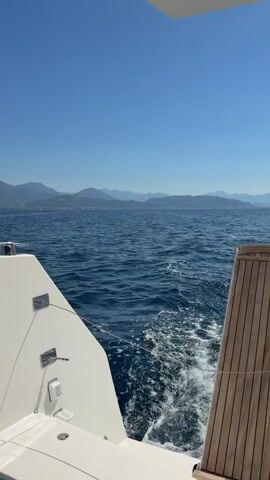 The video shows the back of a white boat, with a wooden panel on the right side. The boat is moving through the blue sea, leaving a white wake. In the distance, there are mountains under a clear blue sky.