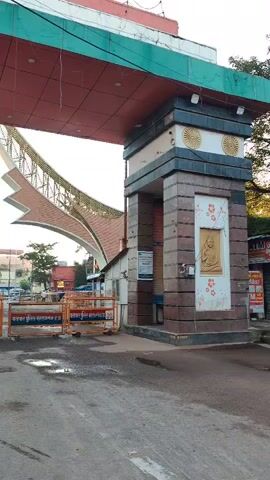 The video shows an outdoor scene at what appears to be a checkpoint or entrance. In the foreground, there is a paved surface with puddles of water, suggesting recent rain. A large, ornate archway dominates the frame, with a red tiled roof and decorative metallic elements. To the right of the archway is a pillar with stone cladding and a decorative panel featuring a Buddha-like figure. Orange traffic cones and police barriers are set up, indicating a controlled entry point. Beyond the barriers, a street scene is visible with buildings, vehicles, and people. The lighting suggests it is either early morning or late afternoon. There are no specific actions or interactions clearly depicted, the focus is on the static environment.