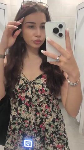 A young woman with long, dark, wavy hair is taking a selfie in what appears to be a bathroom or dressing room. She is wearing a floral print dress and has sunglasses perched on top of her head. She is also wearing a ring on her left ring finger and a bracelet on her right wrist. The phone she is using to take the selfie is white and has three cameras on the back. The background is a tiled wall and a doorframe.
