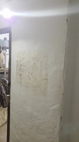 The video showcases a room that appears to be a cellar or storage area. The walls are white and textured, and there is writing in what looks like pencil on them. The ceiling is low and exposed with wooden beams. The room contains several items of old or antique furniture. In the initial view, a glimpse through a doorway reveals an old saddle atop a covered barrel and a woven basket on a shelf. The camera then pans back to reveal an assortment of antique chests, including a green one and a yellow and black one, as well as large wooden tubs, scattered around the room. The door is made of wood and has a metal grate. The lighting is dim, suggesting that the room is either underground or does not have windows.
