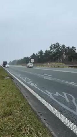 The video shows a shot alongside a highway in what appears to be Europe. Cars pass by on the highway. A black semi-truck hauling what appears to be parts of a parking garage drives past the camera. The weather is overcast and looks damp. The scene is on a highway shoulder with a grassy patch beside the road. There are trees and a sign visible on the other side of the highway.