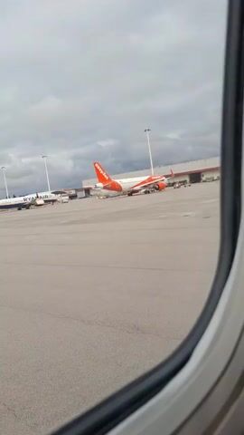The video shows a view out of an airplane window at an airport. In the foreground is the airplane window. In the background are two airplanes parked on the tarmac. On the left is a Ryanair plane. On the right is an EasyJet plane. There are airport service vehicles around the planes. The sky is overcast.