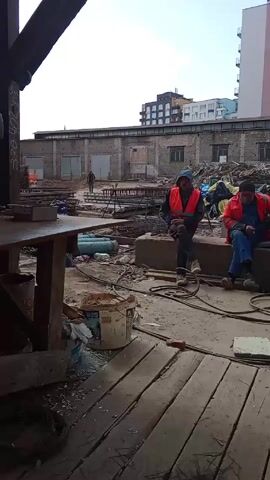 The video shows a construction site, viewed from underneath a wooden structure, where three men in safety vests are sitting on a concrete block. They appear to be on a break, with two of them using their phones and the third smoking a cigarette. The environment is cluttered with construction materials like rebar, hoses, and buckets. There are several low-rise buildings in the background and a taller apartment building further away. A lone figure can be seen walking in the background of the site.
