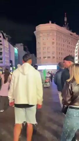 The video shows a nighttime scene in a busy urban area. The camera moves through a crowd of people on a well-lit street. The focus shifts to a tall, white obelisk, likely a monument or landmark. Buildings surround the area, some illuminated with colorful lights. People are walking around, seemingly enjoying the lively atmosphere of the city at night.