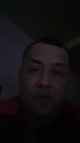 A dimly lit video shows a man looking directly at the camera. He is wearing a dark garment with a red collar. He raises his hand briefly before putting it down. He says "Assalamualaikum" and "Salam semua".