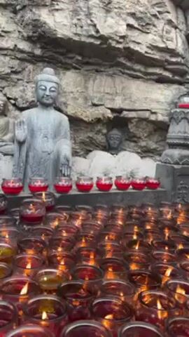 The camera is positioned at eye level and focuses on a collection of lit candles arranged in a grid. The candles are in clear glass containers with red wax. Behind the candles are three carved statues of Buddha set against a rocky, grey wall. The statue on the left is standing with its right hand raised. The statue in the center is reclining. The statue on the right is partially obscured by a stone pillar. The video is well lit and the flames of the candles are clearly visible. The scene appears to be part of a Buddhist temple or shrine. Sounds of people talking and music are present in the audio.
