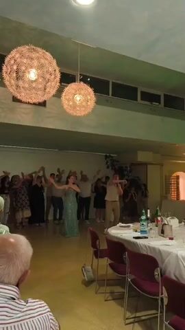 A wide-angle, slightly shaky video shows an indoor event space. Two large, spherical, textured light fixtures hang from the ceiling, illuminating the scene. In the background, a group of people stand and dance, with a woman in a shimmering green dress and a man holding a microphone in the center. The lighting in the room changes colors, cycling through pink, purple, green, and white. In the foreground, a table covered with a white tablecloth is set with plates, glasses, and bottles. The back of an elderly man's head and shoulders, wearing a striped shirt, are visible in the bottom left corner of the frame.