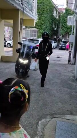 A person in a black helmet and face mask, wearing a black vest over a black long-sleeved shirt and black pants, walks towards the camera while holding a plastic bag in their left hand and a smartphone in their right hand. The person appears to be a delivery person. In the foreground, the back of a young girl's head is visible, with her hair styled in two buns with colorful accessories. Behind the delivery person, a motorcycle with its headlights on is parked. The environment appears to be an alleyway between buildings, with a building covered in ivy on the right side and a building with balconies on the left. Cars are visible in the background.