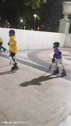 The video shows a group of young people playing roller hockey in an outdoor plaza at night. They are wearing protective gear, including helmets and pads, and using hockey sticks. The plaza appears to be paved with stone and has some architectural features, including columns and stairs, in the background. The lighting suggests it is artificial, likely from street lamps. The action shows two individuals in the foreground engaged in the game, with others further back. The overall scene depicts a recreational activity taking place in a public space after dark.