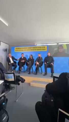 The video shows a panel discussion with five people on a stage. The panel members consist of four people seated on chairs and one person on a video call displayed on a screen in the background. The panel members include three women and two men. The topic of the discussion is visible in a graphic displayed on the stage backdrop and relates to defense technology partnerships, Ukraine, and European security. An audience is viewing the panel discussion from off-camera. At one point, a photographer walks into view to take pictures of the panel.
