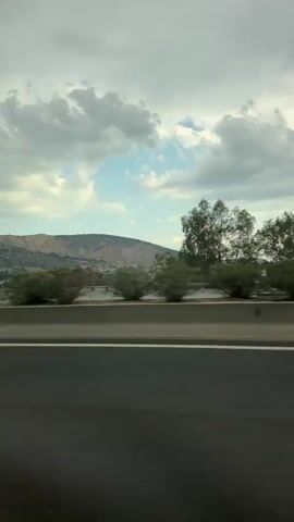 The video shows a view from a moving vehicle on a highway. The foreground consists of the highway lane with white lines. Beyond the highway, there is a median with low green bushes and some trees. In the background, there are hills and mountains, with more trees and vegetation. The sky is cloudy with patches of blue sky visible. The video is shot from a side-facing perspective, indicating the vehicle is traveling forward. There are no people visible in the video. The overall scene suggests a landscape viewed during a car journey.