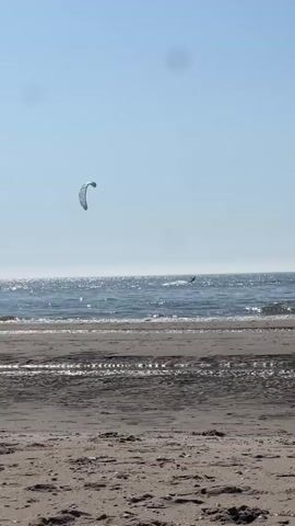 This is a video of a kitesurfer on a beach. The scene is set on a sunny day with a clear blue sky. The ocean is visible in the background with gentle waves, and the sun is glinting off the water, suggesting it is mid-day. The beach in the foreground is sandy, with some debris and tide marks. 

The main subject is a kitesurfer in the distance, on the water. A large, light-colored kite is visible in the air above the surfer, catching the wind. The surfer appears to be actively engaged in the sport, navigating the waves. Another, smaller kite is also visible in the distance, suggesting there might be more than one kitesurfer. 

The overall mood is peaceful and recreational. The clear weather and bright sunlight evoke a sense of a pleasant day at the beach. The location cues, such as the sandy beach and the open ocean, are typical of a coastal environment. There is no visible text in the video.