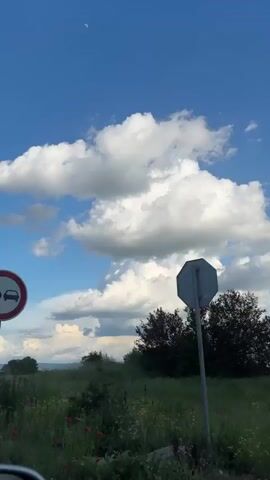 The video shows a roadside scene. First, a faded yellow billboard advertising a hydro park, written in Bulgarian, is visible against a bright blue sky filled with white clouds. The lower part of the billboard pole is defaced with graffiti. The surrounding landscape includes green fields dotted with red poppies. The camera then pans to the right, showcasing a landscape of fields and low-lying trees under the expansive blue sky. As the camera continues to move, it passes by a dense row of trees, creating a blurry effect of green foliage against the bright sky.