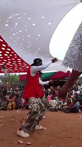 Under a white canopy with red polka dots, two men in matching red basketball jerseys, white long-sleeved shirts, and camouflage pants dance in front of a seated crowd of men, women, and children on a dirt ground. The backdrop is a green and red tent-like structure. The men make arm and leg movements while facing the audience.