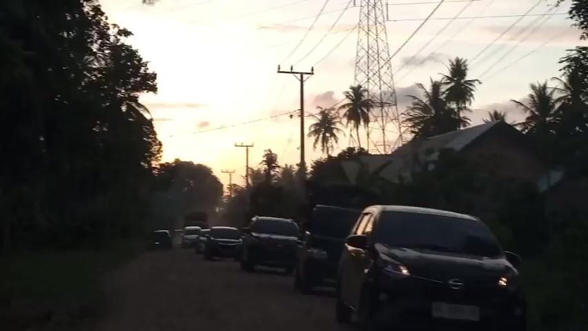 The video shows a dusty, unpaved road lined with trees and palm trees. The sky is overcast with the sun setting in the background, casting a warm glow. Several cars and a truck are on the road, appearing to be in a traffic jam or moving slowly. The cars are dark in color and appear to be SUVs and sedans. There are also some power lines and poles visible along the road. The overall atmosphere is rural and somewhat dusty. The audio consists of ambient sounds and muffled voices, possibly from a conversation or commentary related to the scene.