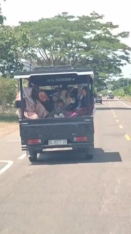 The video shows a rear view of a black pickup truck driving down a paved road. The truck is carrying many people, including women and children, in its open bed. Some of the people are wearing hijabs. The truck has a metal roof over the back. The license plate reads "BL 8177 ZE". There are trees and vegetation on the sides of the road, and the sky is visible. Another white vehicle is driving ahead of the pickup truck. The overall impression is of a crowded vehicle transporting people in a rural or semi-rural setting.