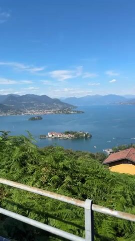 This video shows a panoramic view of a large lake with mountains in the distance. In the foreground, there is lush green foliage and a metal railing. In the middle ground, there is a small island with buildings and trees. The island appears to be inhabited or a tourist destination. The lake is calm and blue, with a few boats visible on the water. The sky is clear and blue with a few scattered clouds. The video also shows a town or city built along the shore of the lake in the distance. The overall impression is one of a beautiful and serene natural landscape with human settlement.