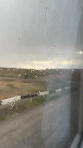 This is a video taken from inside a vehicle, looking out a rain-streaked window at a desolate landscape. The sky is overcast and gray, with some hints of light on the horizon. The ground is a mix of brown and green, with sparse vegetation. There are some scattered bushes and a small tree with silvery leaves. A guardrail runs along the side of the road. The overall impression is one of a gloomy and somewhat empty environment, with the rain adding to the somber mood.