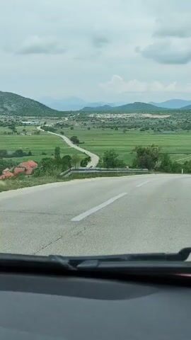 The video shows a view from the interior of a vehicle traveling on a paved road. The road has white lane markings and a metal guardrail on the side. The view from the car is of a green valley with fields and scattered trees. There are mountains visible in the distance under a cloudy sky.