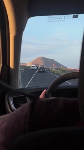 A stationary, interior shot looks through a car windshield at a road with several vehicles in front. A dark gray or black mountain with sparse vegetation rises in the background under a mostly clear, blue sky. A road sign warning of pedestrians ahead is beside the road. The hands of a driver, seen from behind, are on the steering wheel. The driver’s clothing is maroon. The windshield has a small sticker identifying the audio guide and the Canary Islands.