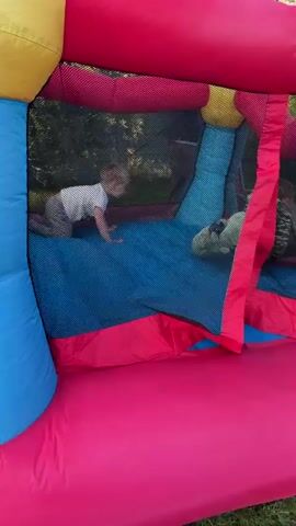 The video shows three young children playing inside a colorful inflatable bounce house. The bounce house is predominantly pink, red, and blue, with yellow accents. The children are visible through a black mesh netting that encloses the play area.

In the foreground, a child wearing a camouflage-patterned jacket and light-colored pants is on their hands and knees, looking towards the camera. To the left, another child in a white shirt and light-colored pants is crawling forward. In the background, a third child, partially obscured, appears to be in a similar crawling or kneeling position.

The children are actively engaged in play, with one child shown in mid-jump in a subsequent frame. The environment outside the bounce house, visible through the netting, suggests an outdoor setting with greenery and trees. The lighting indicates daylight.