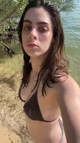 A young woman with wet dark hair, wearing a brown bikini, is standing on a sandy beach.  She is facing the camera and holding it at arm's length. The background includes water, vegetation, and rocks. As the video progresses, the camera moves slightly as she walks, capturing more of the beach and the surrounding environment. The light is bright and sunny.