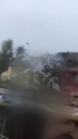 The video shows a blurry view through a window covered in water droplets, likely from rain. Outside, a blurry residential area can be seen, with houses and a tree visible in the distance under a cloudy sky. The focus is on the water on the window, obscuring the view beyond.