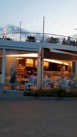 The video pans across an outdoor dining area at dusk. There are several tables with people dining. Some tables are covered by awnings. Lights are strung across the top of the area. In the foreground, people are walking along a paved walkway. The sky is a pale blue with some clouds.