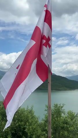 A Georgian flag flutters in the wind on a wooden pole against a backdrop of a vast lake and rolling green hills under a cloudy sky. The flag, predominantly white with red crosses, is positioned in the foreground, partially obscuring the panoramic view. Lush green foliage is visible at the bottom of the frame. The camera remains stationary throughout the video.