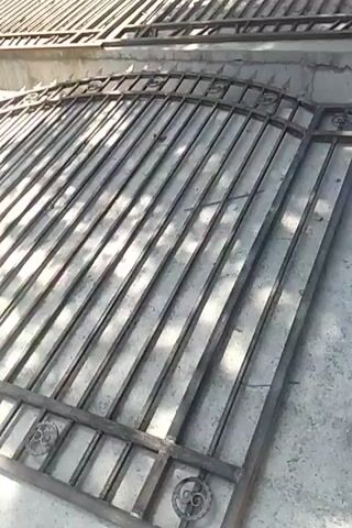This is a close-up, high-angle shot of several metal gates lying on a concrete surface. The gates appear to be unfinished or in storage, with a matte, metallic finish. Each gate is composed of numerous vertical metal bars, evenly spaced, supporting a decorative top edge. The top edge of the gates has an arched design and is adorned with circular metal embellishments, each containing a spiral pattern. The gates are laid out in a row, with some overlapping. The lighting suggests an outdoor setting, with shadows cast across the gates. The concrete surface beneath the gates is rough and appears somewhat weathered.