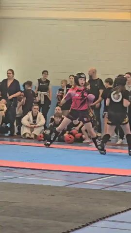 The video shows two fighters, both wearing protective headgear and shin guards, engaged in a kickboxing match on a blue and red mat. The fighter on the left is wearing a black top and shorts with gold trim, while the fighter on the right has a pink and black outfit. Spectators, including adults and children, surround the ring, observing the match. Some are seated on chairs while others are standing. The scene takes place indoors, in what appears to be a gymnasium or community center, with light-colored walls and an overhead lighting system. A white folding chair is visible in the foreground. The match seems to be overseen by a referee.