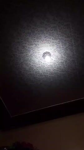A video shows a ceiling with a light fixture in the center. The ceiling has a patterned design and is dark around the edges. The light fixture is illuminated and casts a bright glow. The camera angle is from below, looking up at the ceiling.
