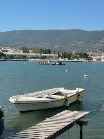 The video captures a serene waterfront scene on a clear, sunny day in Poros, Greece. The camera slowly pans from left to right, showcasing the tranquil bay and its surroundings.

In the foreground, a small white dinghy with a yellow stripe is partially visible on the left, moored near a wooden dock. Text "Π.Λ. 11982 A" is visible on its hull. As the camera pans, a larger white motorboat comes into full view, tied to the wooden dock. This boat has a white canopy over its cockpit and a Greek flag fluttering at its stern. The name "ΣΕΡΑΦΙΝΟ Λ.Κ. 2830" is clearly visible on its bow.

The calm, blue-green water of the bay is dotted with a few buoys and other distant boats. Across the water, a charming town or village with white and light-colored buildings is nestled at the foot of rolling green mountains. Several white wind turbines are visible along the ridge of the distant mountains under a bright blue sky. The overall impression is one of peaceful coastal scenery.