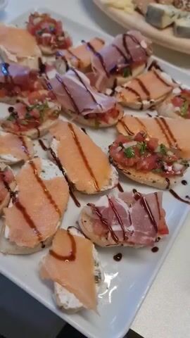 The video showcases a platter of various bruschetta appetizers. The bruschetta are topped with different ingredients, including chopped tomatoes, smoked salmon, and prosciutto. Some pieces are drizzled with a dark sauce, likely balsamic glaze. The appetizers are arranged on a white rectangular platter. In the background, another platter of similar appetizers is partially visible. The setting appears to be indoors, possibly a restaurant or a catered event, with a neutral background. The lighting suggests it is daytime. There is no visible text in the video. The focus is entirely on the food presentation, which is neat and appealing.