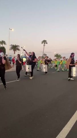 The video shows a parade moving down a street at twilight. A group of people dressed in black shirts and pants, and wearing drums, are walking and playing the drums. In front of them are people dressed in green and black costumes with tall, elaborately decorated hats. Some palm trees are visible, as well as other people watching the parade. The sky is pale blue.