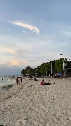 A wide shot of a sandy beach with several people enjoying the late afternoon sun. The sky is a soft blue with scattered clouds, some tinged with orange and pink from the setting sun. In the foreground, the sand is littered with small pieces of debris. Further back, a group of people are walking along the water's edge, while others are relaxing on the sand. Some are lying down, perhaps sunbathing or resting. Further still, there are several small stalls and buildings, suggesting a beachfront community or marketplace. Trees line the back of the beach, providing a green backdrop. The overall atmosphere is peaceful and relaxed.