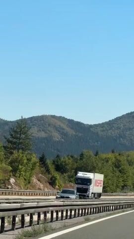 The video shows a view from a car driving on a highway. The highway has multiple lanes and is bordered by a metal guardrail. On the right side of the highway, there is a steep, rocky incline covered with trees. The landscape is mountainous and covered with dense green forests. The sky is clear and blue. A white truck with a red logo that says "MISPED" is visible on the highway. Several other cars are also on the road. The sun is shining, indicating it is daytime. The camera appears to be mounted inside a vehicle, capturing the road and scenery as it moves.