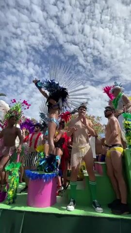 The video displays a lively outdoor scene with several individuals dressed in vibrant, feathered costumes on a green platform, likely a parade float. The sky above is a bright blue with scattered white clouds. The people are adorned with elaborate headdresses, colorful feathers, and minimal clothing, including briefs and decorative mesh garments. One person stands on a pink pedestal, arms raised, wearing a large, silver and blue feathered headdress. Another person in the foreground wears a sheer, netted garment over yellow briefs and green socks. The overall atmosphere appears festive, possibly during a carnival or pride event. Palm trees are visible in the background, suggesting a warm climate.
