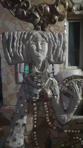 A statue of a woman is shown from the chest up. The statue is covered in mosaic tiles of various colors and textures. The woman has a stern expression and her hair is styled in a blocky fashion. She is wearing a mosaic necklace and has a mosaic dress. Her right arm is raised and she appears to be holding something in her hand, possibly a fan or a flower. The background is blurred but appears to be an outdoor setting with some architectural elements visible. The lighting is dim and the overall mood is somewhat mysterious.