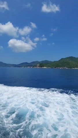The video shows the view from a boat, with the ocean in the foreground and an island in the background. The water is deep blue, with white foamy waves being created by the boat's movement. The island is green with dense vegetation and some buildings visible along the coastline. The sky is a clear blue with a few scattered white clouds.