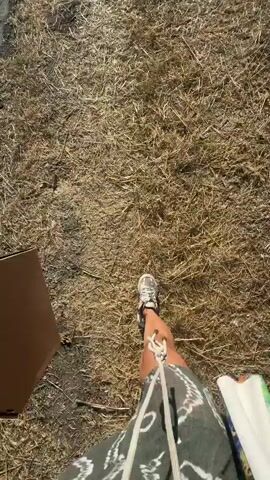 The video shows a first-person perspective of someone walking on a dry, grassy surface. The person is wearing sneakers and a patterned skirt. They are carrying a large, flat, brown object in their left hand and a colorful bag or item in their right hand. The sun appears to be shining, casting shadows. The ground is covered in dry, straw-like grass.