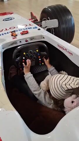 A child wearing a striped beanie and a grey jacket sits in a toy race car, holding a black steering wheel. The child is leaning back, and a stuffed animal is visible behind them. The toy car is white with red accents and has the words "Toyota F1" and "EMC" printed on it. The background appears to be indoors with a light-colored floor.