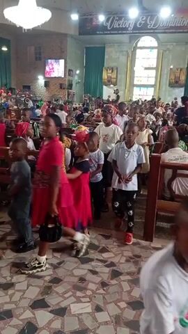 The video shows the interior of a church filled with people, primarily children. Most children are dressed in casual clothing and some are wearing red. The adults are seated in the pews while the children move around. The church has high ceilings, a chandelier, and large windows. There is a stage area at the front with people in red and white robes. The floor is patterned with tiles. Some people are walking around. The overall atmosphere seems lively and possibly celebratory.
