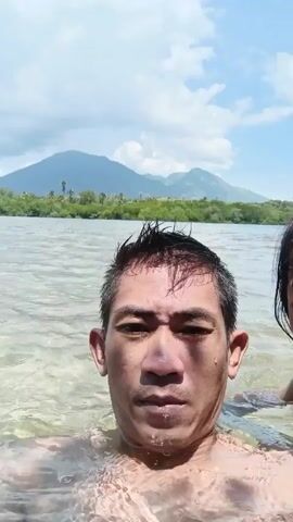 Here is a brief, forensic description of the video:

The video depicts a man and a young girl, waist-deep in shallow, slightly murky water. The man appears to be holding the camera, presenting a selfie-style view. The girl is positioned slightly behind and to the side of the man, partly visible in the frame. Both subjects have wet hair. In the background, across the water, there is land covered in green vegetation and a distinct mountain range. The sky is blue with scattered clouds. The subjects are still for most of the video, however, some utterances are made throughout the shot.
