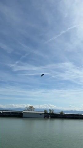 This video shows a blimp flying in a blue sky with scattered clouds. The blimp has "ZF Next Generation Mobility" and "NOW" written on its side in white letters. There is also a smaller airplane visible in the distance. The blimp is in the center of the frame, with the sky filling most of the screen. The bottom of the frame shows a body of water and a pier with a building on it.