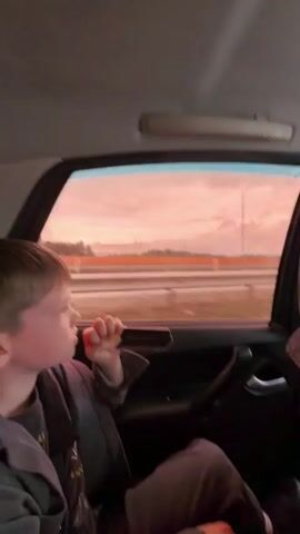 A young child, seen from the side, sits in the back seat of a car, looking out the window. The child is holding a small, dark object to their mouth, appearing to suck on it. The car's interior is visible, with part of a car door, handle, and a seat headrest. Outside the tinted window, a road with multiple lanes is visible, separated by a barrier. The sky is a soft pinkish-orange, suggesting either sunrise or sunset, with some clouds and structures, possibly industrial, in the distance. The overall scene depicts a moment of quiet observation during a car journey.